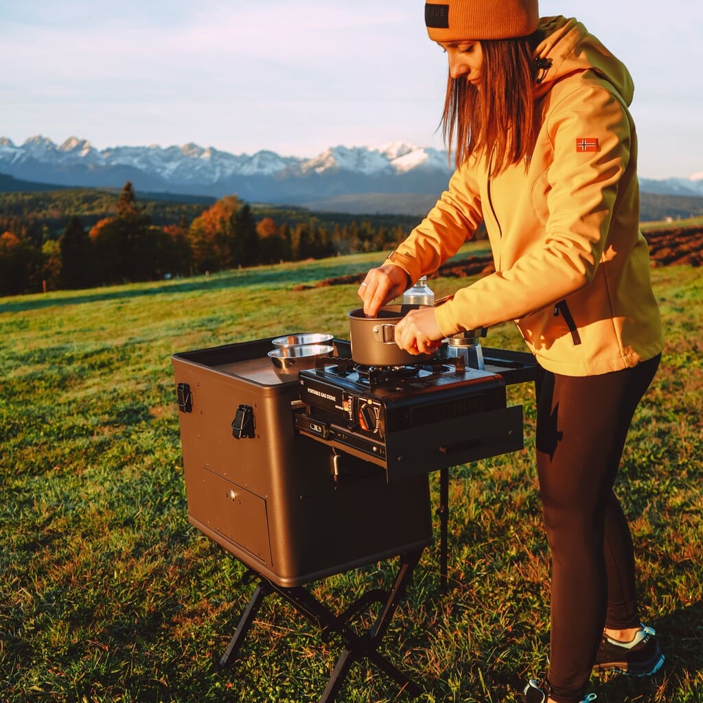 Campingküchenbox auf Wiese im Herbst bei Sonnenschein. Frau kocht auf Gaskocher auf der LAYZEE Kitchenbox.
